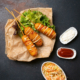 Drei Corndogs liegen auf einem Stück Backpapier, daneben drei kleine Schälchen mit Ketchup, Mayonnaise und Coleslaw