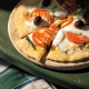 Eine Erdäpfelpizza belegt mit Tomaten, Käse und Oliven auf einem runden Holzbrett