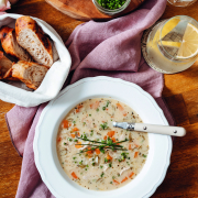 Ein gedeckter Tisch, auf dem ein Brotkörbchen, in Röllchen geschnittener Schnittlauch, ein Glas mit einem Getränk, sowie ein weißer Teller mit Suppe angerichtet sind