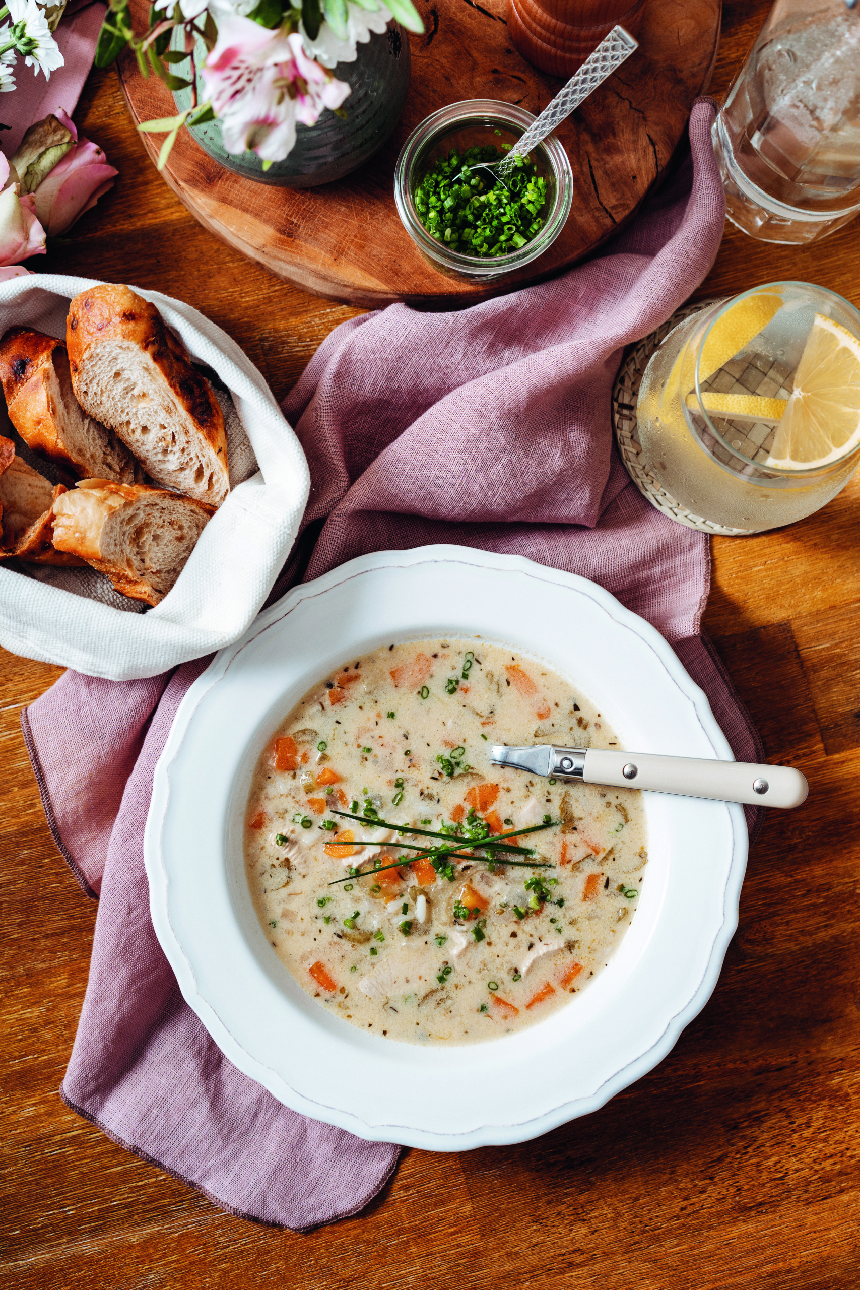 Ein gedeckter Tisch, auf dem ein Brotkörbchen, in Röllchen geschnittener Schnittlauch, ein Glas mit einem Getränk, sowie ein weißer Teller mit Suppe angerichtet sind