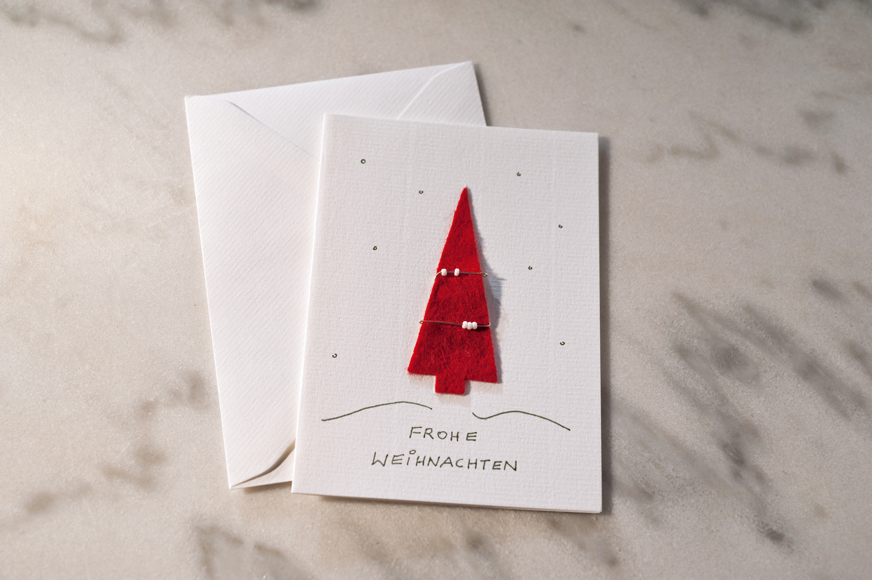 Ein weißes Billet mit einem roten Filztannenbaum, der mit auf Draht aufgefädelten Perlen dekoriert ist. Darunter der Schriftzug Frohe Weihnachten