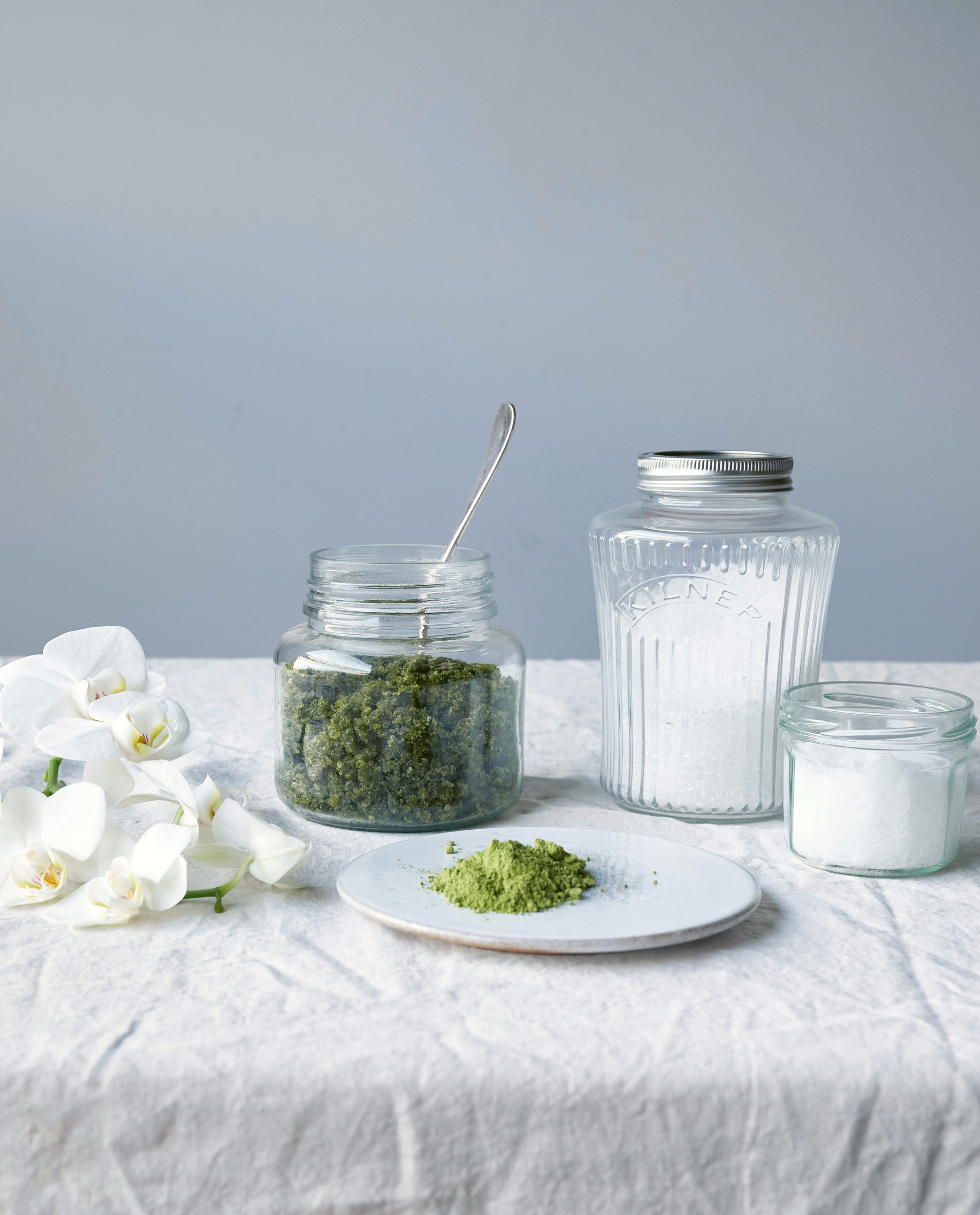 Drei Glasbehälter gefüllt mit Badesalz auf einem weißen Tischtuch, daneben eine Orchidee als Deko