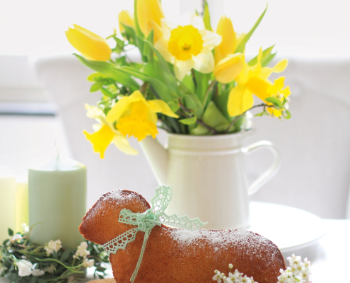 Ein Kuchen in Form eines Osterlamms auf einem Holzbrett, das auf einem frühlingshaft dekorierten Tisch steht
