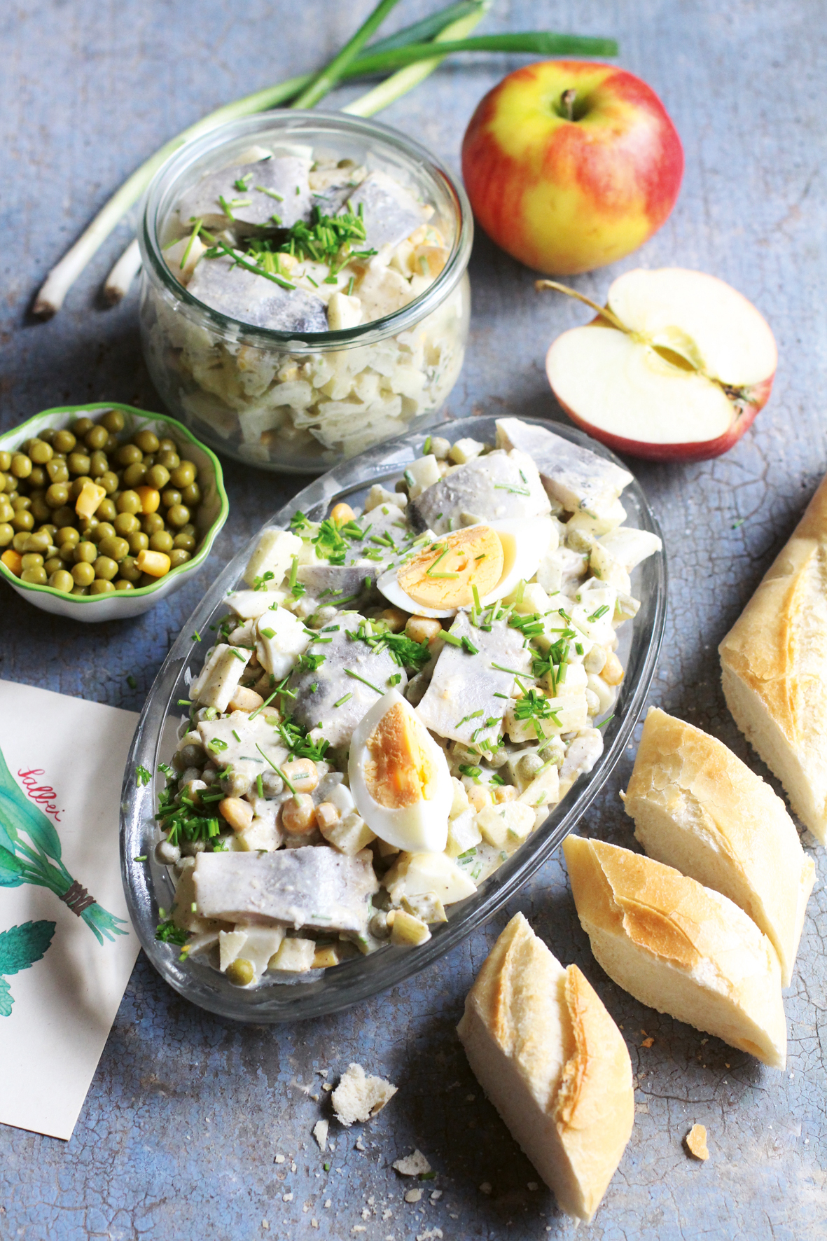 Heringsalat garniert mit Ei und Schnittlauch in einer Glasschale. Rund um die Schale befindet sich ein Apfel, ein Baguette und noch ein volles Glas mit Heringsalat.