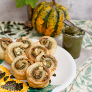 Ein Teller auf dem gerollte Blätterteigschnecken mit Kürbiskernpesto als Fülle liegen, daneben steht ein Glas mit Kürbiskernpesto und ein Kürbis.