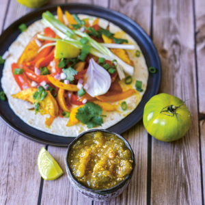 Auf einem Teller liegt ein Taco mit Gemüse, davor steht eine Schüssel mit grüner Soße bzw. Salsa Verde.