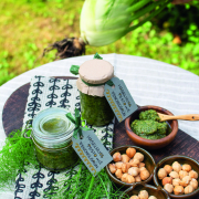Auf einem Gartentisch liegt ein dunkles Holzbrette, auf dem zwei Gläser mit Haselnusspesto stehen, daneben sind vier kleine Schüsseln, drei sind gefüllt mit Haselnüsse, eines mit Pesto.