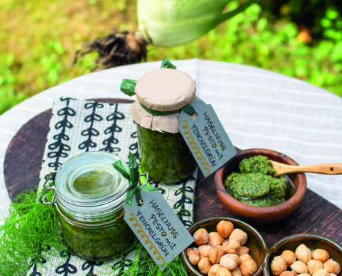 Auf einem Gartentisch liegt ein dunkles Holzbrette, auf dem zwei Gläser mit Haselnusspesto stehen, daneben sind vier kleine Schüsseln, drei sind gefüllt mit Haselnüsse, eines mit Pesto.