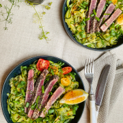 Zwei Teller auf dem jeweils das aufgeschnittene Thunfischsteak auf den Zucchininudeln angerichtet wurde. Garniert mit Tomaten und einer Zitronenspalte. Daneben liegen Gabel und Messer auf einer zusammengefalteten Serviette.