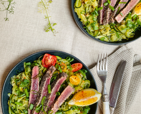 Zwei Teller auf dem jeweils das aufgeschnittene Thunfischsteak auf den Zucchininudeln angerichtet wurde. Garniert mit Tomaten und einer Zitronenspalte. Daneben liegen Gabel und Messer auf einer zusammengefalteten Serviette.