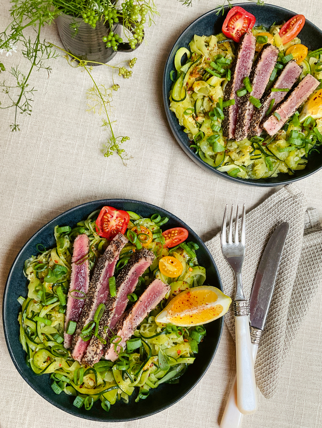 Zwei Teller auf dem jeweils das aufgeschnittene Thunfischsteak auf den Zucchininudeln angerichtet wurde. Garniert mit Tomaten und einer Zitronenspalte. Daneben liegen Gabel und Messer auf einer zusammengefalteten Serviette.