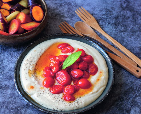 Ein dunkler Teller auf dem die Aioli angerichtet ist, darauf sind Tomaten platziert. Neben dem Teller liegt ein Holzbesteck