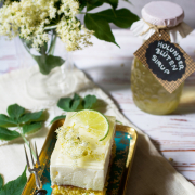 Ein Stück Holunderblütenschnitte mit Limettenscheibe und frischen Holunderblüten auf einem Teller, daneben ein Glas Holunderblütensirup und ein Strauß Holunderblüten.