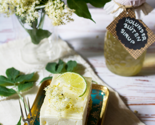 Ein Stück Holunderblütenschnitte mit Limettenscheibe und frischen Holunderblüten auf einem Teller, daneben ein Glas Holunderblütensirup und ein Strauß Holunderblüten.