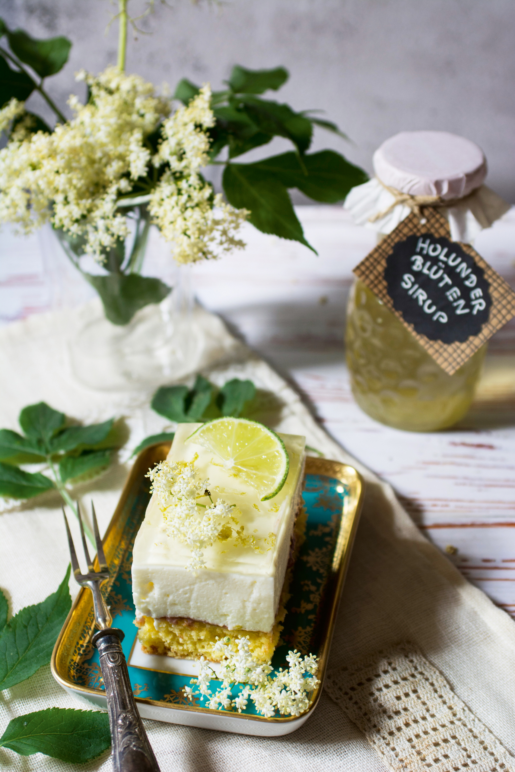 Ein Stück Holunderblütenschnitte mit Limettenscheibe und frischen Holunderblüten auf einem Teller, daneben ein Glas Holunderblütensirup und ein Strauß Holunderblüten.
