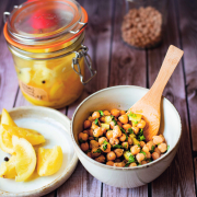 Ein Einmachglas mit eingelegten Zitronen, daneben steht eine kleine Schüssel mit Kichererbsen und einem Holzlöffel. Daneben noch ein Teller, auf dem ein paar eingelegte Zitronenstücke liegen.
