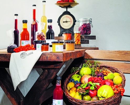 Ein Holztisch, auf dem viele Obstsaftflaschen und Marmeladegläser stehen. Daneben ein Holzsessel, auf dem ein Korb mit frischem Obst und Gemüse steht