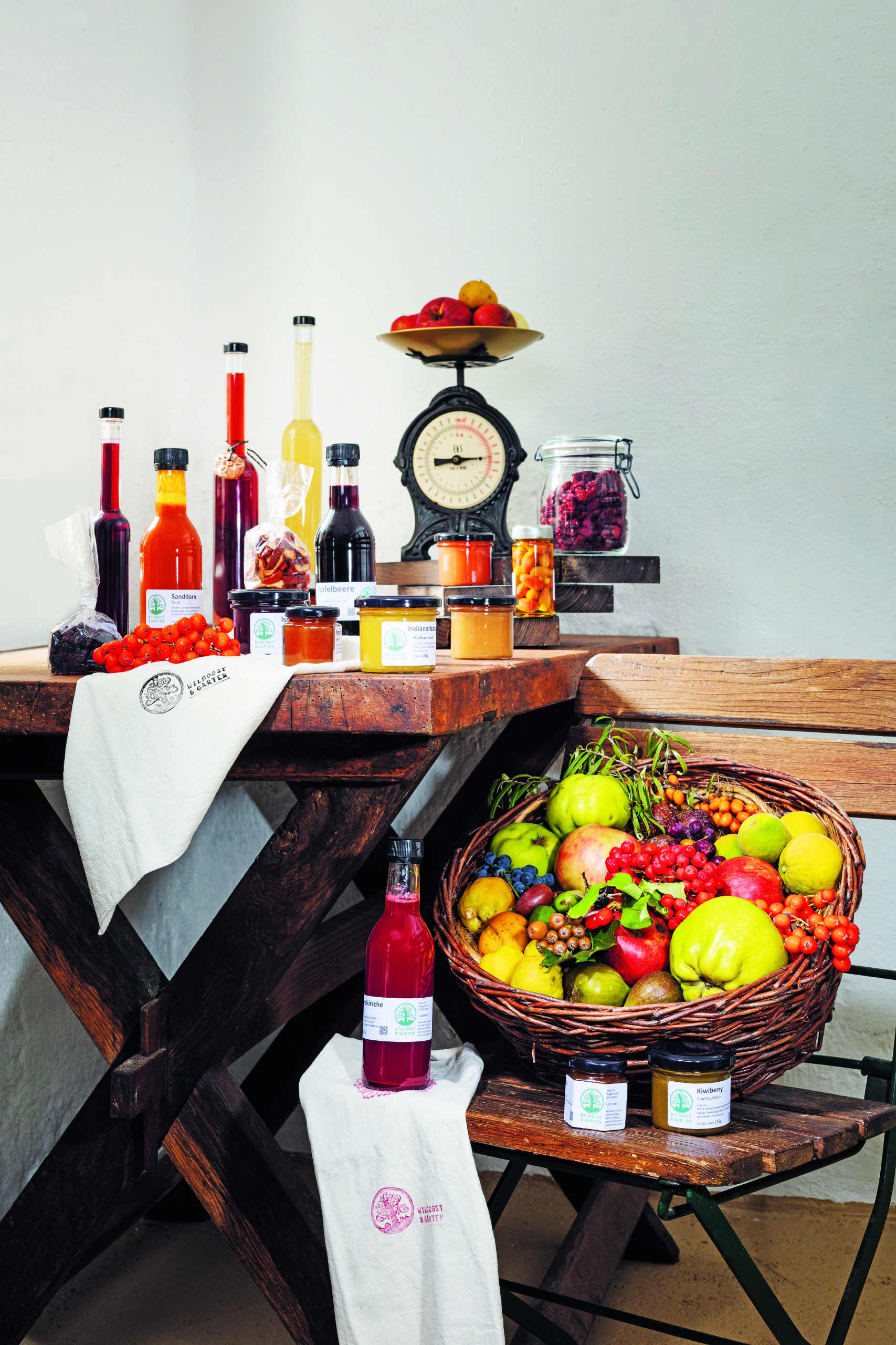 Ein Holztisch, auf dem viele Obstsaftflaschen und Marmeladegläser stehen. Daneben ein Holzsessel, auf dem ein Korb mit frischem Obst und Gemüse steht Ein Holztisch, auf dem viele Obstsaftflaschen und Marmeladegläser stehen. Daneben ein Holzsessel, auf dem ein Korb mit frischem Obst und Gemüse steht