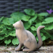 Eine sitzende, aus Holz geschnitzte Katze vor einer grünen Pflanze.