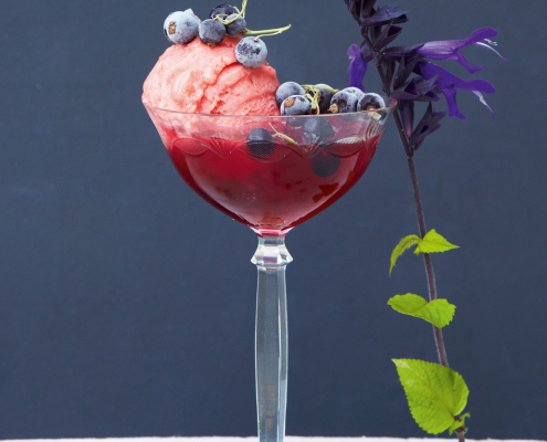 Ein durchsichtiges Cocktailglas, gefüllt mit rotem Salbeiblütensirup, Erdbeersorbet und Heidelbeeren