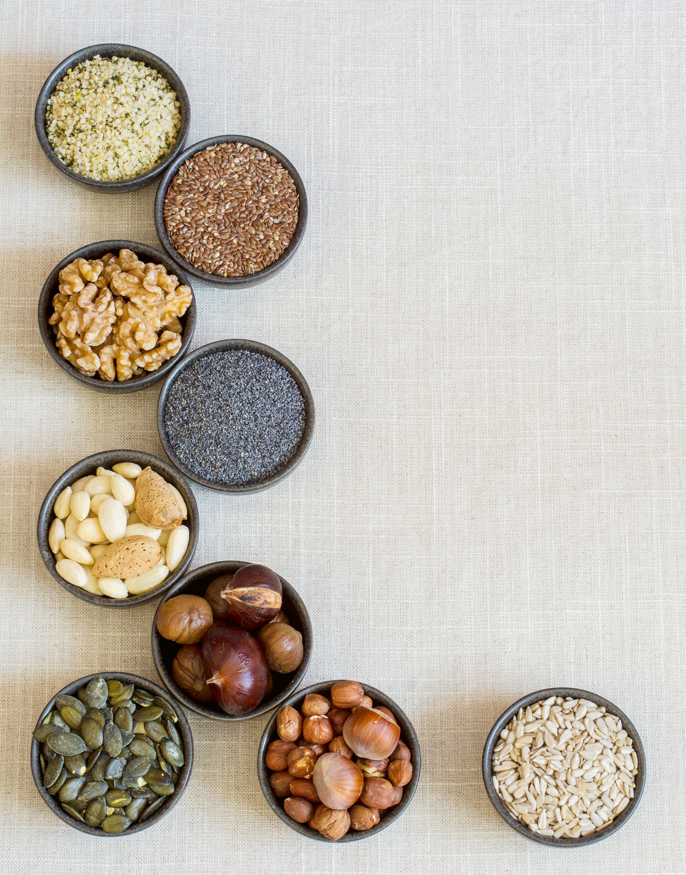 Insgesamt neun schwarze kleine Schälchen, die verschiedene Samen und Nüsse enthalten. Es sind Walnüsse, Mandeln, Haselnüsse, Leinsamen, Hanfsamen, Maroni, Mohn, Sonnenblumen- und Kürbikerne.
