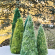 Fünf grüne gefilzte Tannenbäume in verschiedener Größe stehen auf einem Tisch vor einem Nadelbaum.
