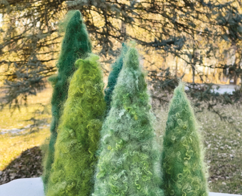 Fünf grüne gefilzte Tannenbäume in verschiedener Größe stehen auf einem Tisch vor einem Nadelbaum.