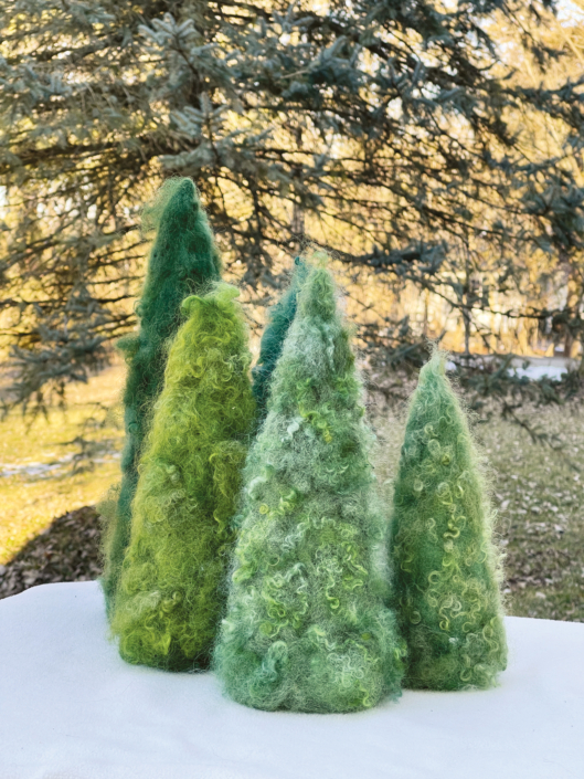 Fünf grüne gefilzte Tannenbäume in verschiedener Größe stehen auf einem Tisch vor einem Nadelbaum.