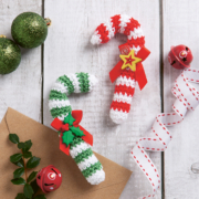 zwei gehäkelte Zuckerstangen, eine in rot-weiß und die andere in grün-weiß liegen auf einem Tisch. Daneben liegen grüne Weihnchachskugeln und rote Glöckchen