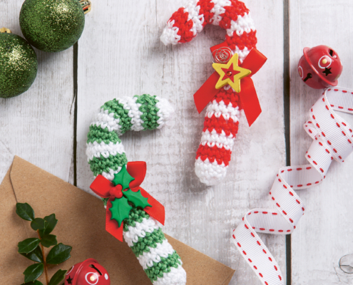 zwei gehäkelte Zuckerstangen, eine in rot-weiß und die andere in grün-weiß liegen auf einem Tisch. Daneben liegen grüne Weihnchachskugeln und rote Glöckchen