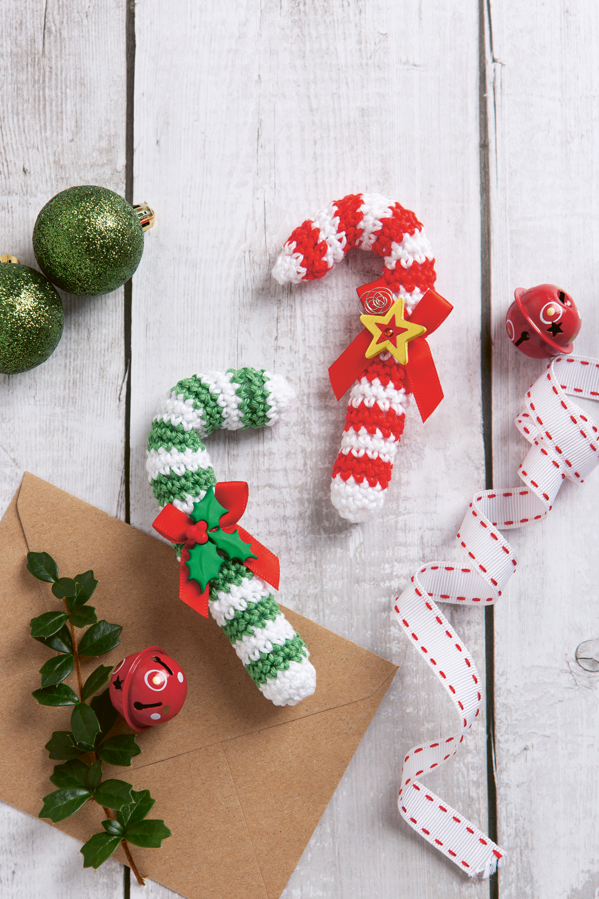 Niedliche Häkel-Ideen f. Weihnachten34_C_Search Press Ltd. 2024 zwei gehäkelte Zuckerstangen, eine in rot-weiß und die andere in grün-weiß liegen auf einem Tisch. Daneben liegen grüne Weihnchachskugeln und rote Glöckchen