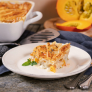 Ein Stück vom herbstlichen Pie liegt auf einem weißen Teller. Daneben die Auflaufform mit der restlichen Pie und ein halbierter Kübis.