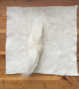 Ein weißes quadratisches Stück Filz auf dem ein Strang Wolle liegt. Ein weißes quadratisches Stück Filz auf dem ein Strang Wolle liegt.