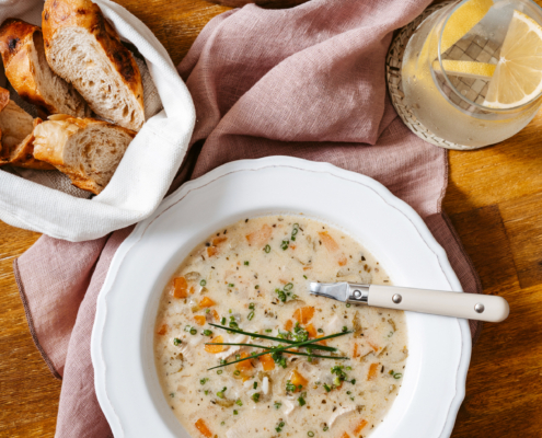 Im Vordergrund steht ein weißer Teller mit Hühneruppe auf einem braunen Holztisch. Dahinter befindet sich ein kleiner Brotkorb, eine kleine Glasschale mit Schnittlauch und ein gefülltes Glas.