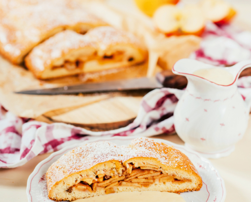 Ein Stück Apfelstrudel mit Vanillesauce ist auf einem Teller mit Löffel und Gabe angerichtet. Daneben steht ein Kännchen und im Hintergrund liegt der restliche Strudel auf einem Holzbrett.