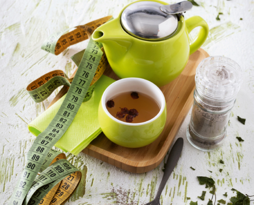 Auf einem braunen rechteckigen Holztablett steht eine kleine grüne Kanne und eine befüllte Teetasse. Daneben liegt ein Maßband sowie Serviette und ein Löffel