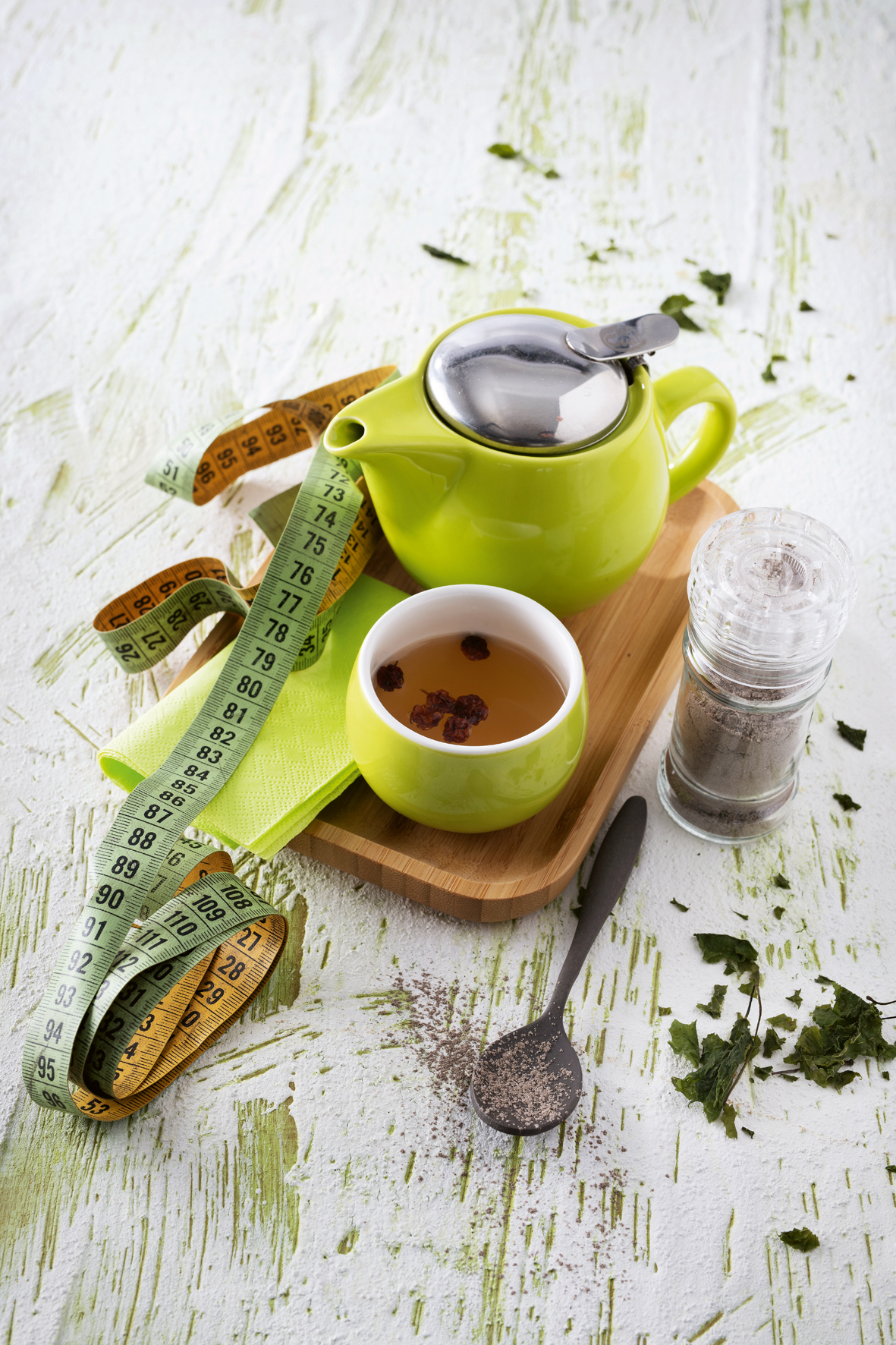 Auf einem braunen rechteckigen Holztablett steht eine kleine grüne Kanne und eine befüllte Teetasse. Daneben liegt ein Maßband sowie Serviette und ein Löffel