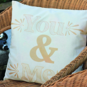 Ein weißes Kissen mit einem beigen Aufdruck You & Me liegt auf einem braunen Korbsessel