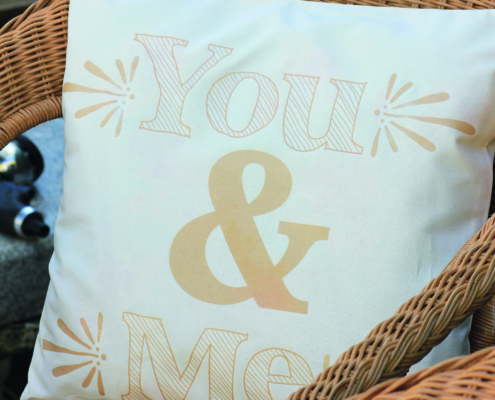Ein weißes Kissen mit einem beigen Aufdruck You & Me liegt auf einem braunen Korbsessel