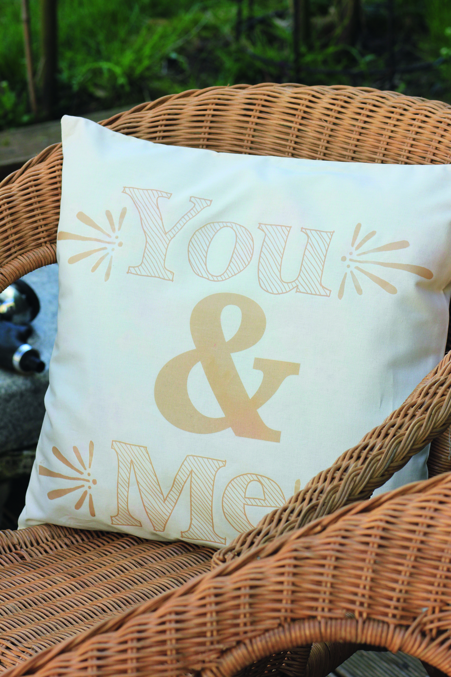 Ein weißes Kissen mit einem beigen Aufdruck You & Me liegt auf einem braunen Korbsessel