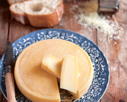Auf einer Holztischplatte steht ein blau-weißer Teller, auf dem ein runder Käse liegt. Davon wurde ein Stück herunterschnitten und liegt oben auf, danben befindet sich ein Messer.