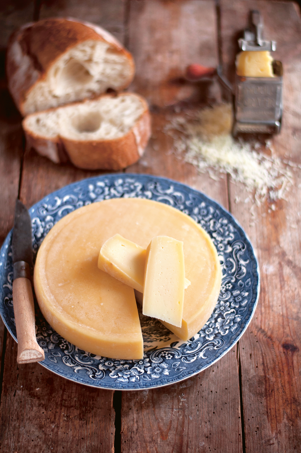 Auf einer Holztischplatte steht ein blau-weißer Teller, auf dem ein runder Käse liegt. Davon wurde ein Stück herunterschnitten und liegt oben auf, danben befindet sich ein Messer.