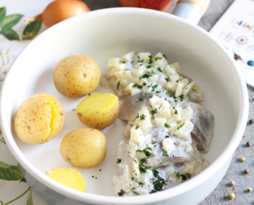 In einer kleinen Pfanne, die auf einem Tisch steht, ist der Hering in Sahnesauce zusammen mit einigen Kartoffeln angerichtet. Im Hintergrund liegen zwei Zwiebeln und ein Apfel als Deko.