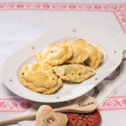 Mehrere Kärntner Kasnudeln sind auf einem weißen ovalen Teller angerichtet. Daneben liegt ein Holzkochlöfel, auf dem Kärnten eingraviert ist.