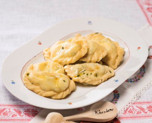 Mehrere Kärntner Kasnudeln sind auf einem weißen ovalen Teller angerichtet. Daneben liegt ein Holzkochlöfel, auf dem Kärnten eingraviert ist.