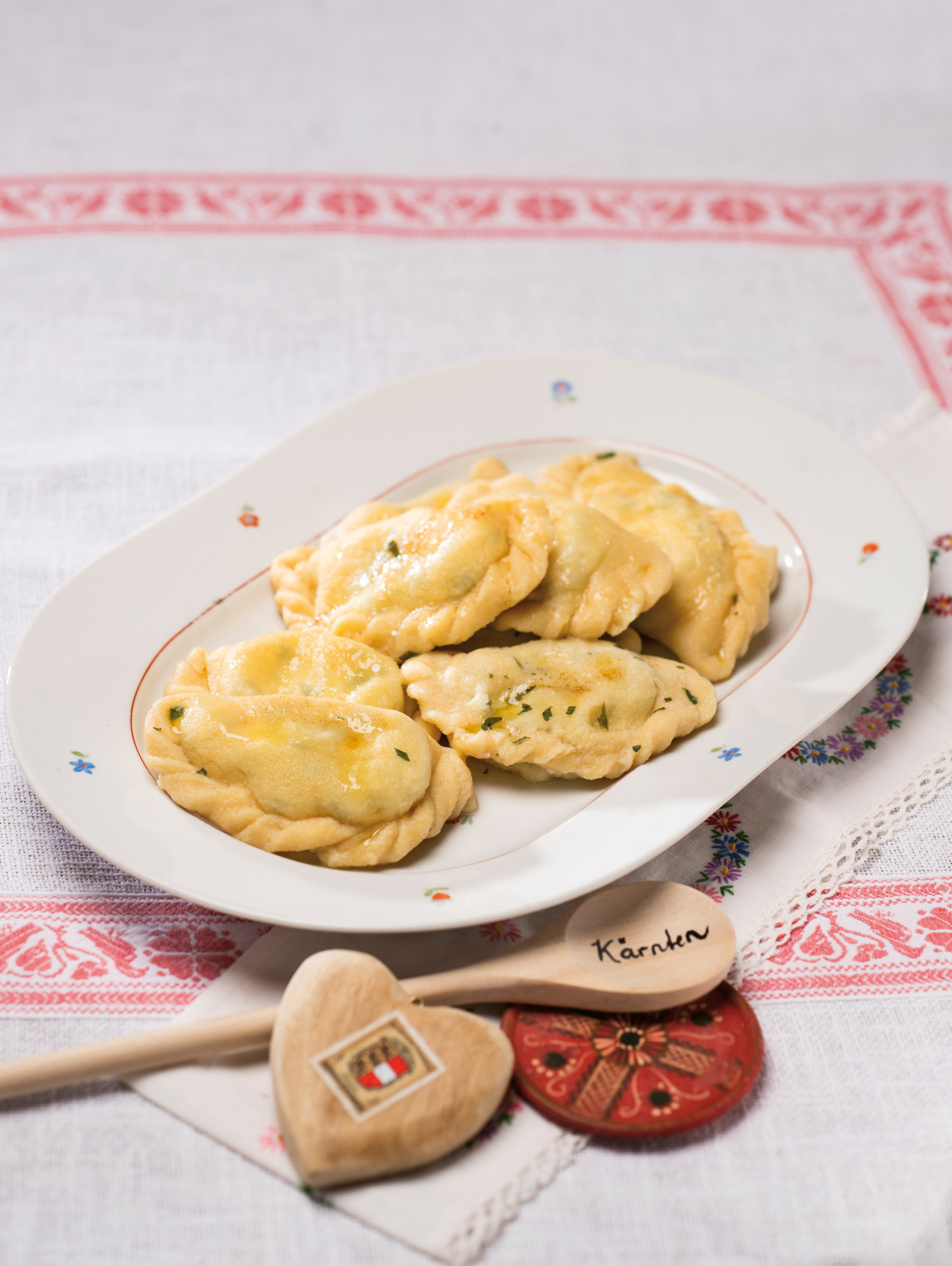 Mehrere Kärntner Kasnudeln sind auf einem weißen ovalen Teller angerichtet. Daneben liegt ein Holzkochlöfel, auf dem Kärnten eingraviert ist.