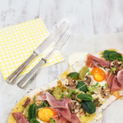 Ein belegter Flammkuchen mit Schinken, Pesto, Champignons, Spinat und Spiegeleier. Daneben liegen Messer und Gabel auf einer gelben Serviette.