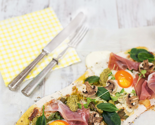 Ein belegter Flammkuchen mit Schinken, Pesto, Champignons, Spinat und Spiegeleier. Daneben liegen Messer und Gabel auf einer gelben Serviette.