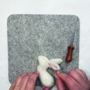 Ein quadratisches Stück Filz liegt auf einem weißen Tisch. Darauf sieht man zwei Hände, die die Ohren des Kaninchens anfilzen.