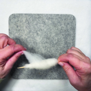 Ein quadratisches Stück Filz liegt auf einem weißen Tisch. Darauf sieht man zwei Hände, die etwas weiße Wolle um ein Holzstäbchen wickeln.